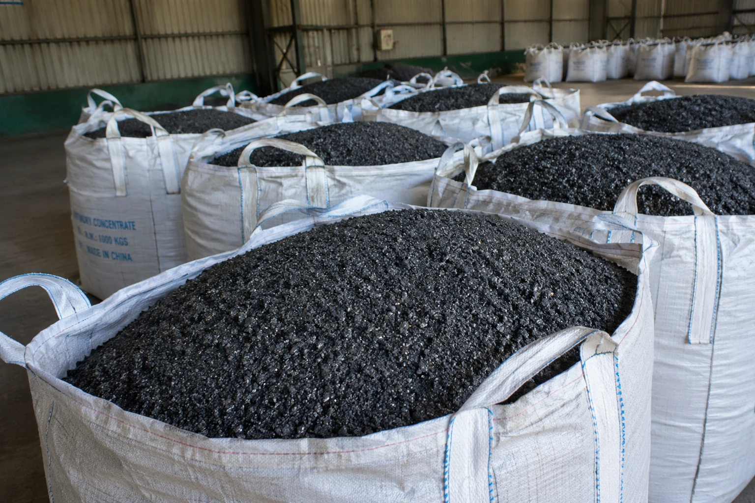 Dark granular antimony concentrate stored in open FIBC bulk bags inside an industrial warehouse